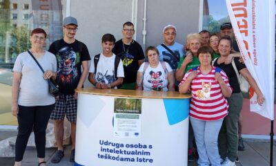 Na fotografiji je skupina članova i djelatnika Udruge osoba s intelektualnim teškoćama Istre okupljena ispred promotivnog štanda tijekom javnog predstavljanja rada Udruge. U prvom planu nalazi se bijeli promotivni pult s natpisom "Udruga osoba s intelektualnim teškoćama Istre", na kojem je postavljen plakat Erasmus+ projekta "Nature & Disabilities". Iza pulta stoji jedanaest osoba različite dobi koje poziraju za zajedničku fotografiju. Većina ih se smiješi prema kameri, a atmosfera djeluje opušteno i prijateljski. S desne strane fotografije vidi se promotivna zastava Udruge, dok se u pozadini nalaze staklene površine zgrade i dio urbanog okruženja. Sunčano vrijeme i vanjski prostor upućuju na to da je fotografija snimljena tijekom javnog događanja ili promotivne akcije. Fotografija prikazuje zajedništvo članova Udruge te naglašava njihov angažman u predstavljanju projekata i aktivnosti kojima se promiče inkluzija osoba s intelektualnim teškoćama.