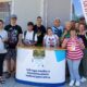 Na fotografiji je skupina članova i djelatnika Udruge osoba s intelektualnim teškoćama Istre okupljena ispred promotivnog štanda tijekom javnog predstavljanja rada Udruge. U prvom planu nalazi se bijeli promotivni pult s natpisom "Udruga osoba s intelektualnim teškoćama Istre", na kojem je postavljen plakat Erasmus+ projekta "Nature & Disabilities". Iza pulta stoji jedanaest osoba različite dobi koje poziraju za zajedničku fotografiju. Većina ih se smiješi prema kameri, a atmosfera djeluje opušteno i prijateljski. S desne strane fotografije vidi se promotivna zastava Udruge, dok se u pozadini nalaze staklene površine zgrade i dio urbanog okruženja. Sunčano vrijeme i vanjski prostor upućuju na to da je fotografija snimljena tijekom javnog događanja ili promotivne akcije. Fotografija prikazuje zajedništvo članova Udruge te naglašava njihov angažman u predstavljanju projekata i aktivnosti kojima se promiče inkluzija osoba s intelektualnim teškoćama.