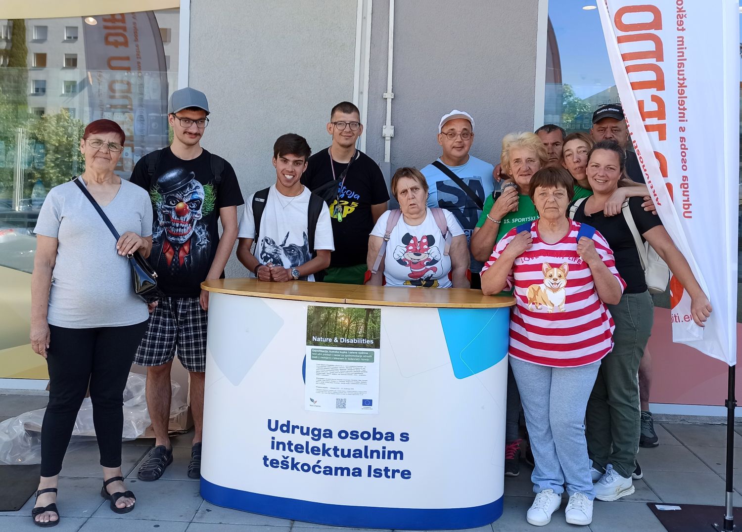 Na fotografiji je skupina članova i djelatnika Udruge osoba s intelektualnim teškoćama Istre okupljena ispred promotivnog štanda tijekom javnog predstavljanja rada Udruge. U prvom planu nalazi se bijeli promotivni pult s natpisom "Udruga osoba s intelektualnim teškoćama Istre", na kojem je postavljen plakat Erasmus+ projekta "Nature & Disabilities". Iza pulta stoji jedanaest osoba različite dobi koje poziraju za zajedničku fotografiju. Većina ih se smiješi prema kameri, a atmosfera djeluje opušteno i prijateljski. S desne strane fotografije vidi se promotivna zastava Udruge, dok se u pozadini nalaze staklene površine zgrade i dio urbanog okruženja. Sunčano vrijeme i vanjski prostor upućuju na to da je fotografija snimljena tijekom javnog događanja ili promotivne akcije. Fotografija prikazuje zajedništvo članova Udruge te naglašava njihov angažman u predstavljanju projekata i aktivnosti kojima se promiče inkluzija osoba s intelektualnim teškoćama.
