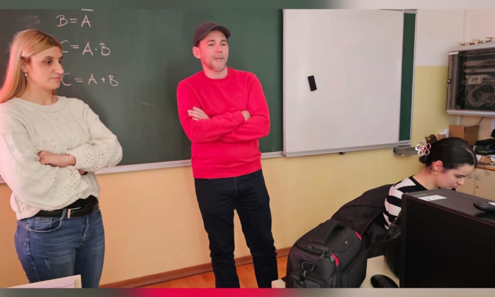 Na fotografiji se nalaze tri osobe u učionici ili informatičkom kabinetu. U središtu fotografije stoji muškarac u crvenoj majici i tamnoj kapi, prekriženih ruku, dok govori ili se obraća prisutnima. S njegove lijeve strane stoji žena u svijetlom džemperu i trapericama, također prekriženih ruku, koja pažljivo prati događanja. S desne strane sjedi žena za računalom i radi na računalu. U pozadini se vidi školska ploča s ispisanim matematičkim formulama i bijela projekcijska ploča, što upućuje na to da je fotografija snimljena tijekom predavanja, radionice ili edukativne aktivnosti u školi. Atmosfera djeluje radno i edukativno, a sudionici su usmjereni na zajedničku aktivnost ili prezentaciju.