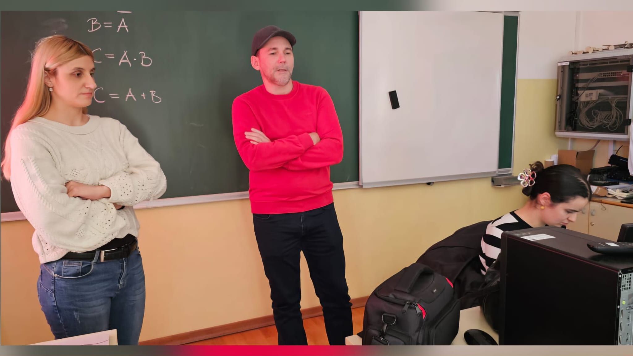 Na fotografiji se nalaze tri osobe u učionici ili informatičkom kabinetu. U središtu fotografije stoji muškarac u crvenoj majici i tamnoj kapi, prekriženih ruku, dok govori ili se obraća prisutnima. S njegove lijeve strane stoji žena u svijetlom džemperu i trapericama, također prekriženih ruku, koja pažljivo prati događanja. S desne strane sjedi žena za računalom i radi na računalu. U pozadini se vidi školska ploča s ispisanim matematičkim formulama i bijela projekcijska ploča, što upućuje na to da je fotografija snimljena tijekom predavanja, radionice ili edukativne aktivnosti u školi. Atmosfera djeluje radno i edukativno, a sudionici su usmjereni na zajedničku aktivnost ili prezentaciju.