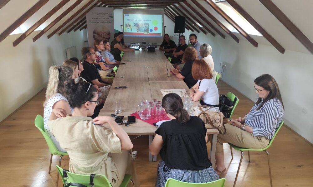 Na fotografiji je prikazan sastanak ili konferencija održana u potkrovnoj dvorani. Oko velikog drvenog stola sjedi petnaestak sudionika koji prate prezentaciju projiciranu na platnu na kraju prostorije. U pozadini se vidi i promotivni roll-up s nazivom projekta „Odmori se, imaš podršku“. Sudionici sjede u opuštenoj, radnoj atmosferi, a na stolu se nalaze čaše, vrčevi s vodom i osobni predmeti, što upućuje na radni sastanak ili završnu konferenciju projekta. Prostorija je osvijetljena prirodnim svjetlom koje dolazi kroz krovne prozore, stvarajući ugodnu i profesionalnu atmosferu.