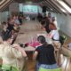 Na fotografiji je prikazan sastanak ili konferencija održana u potkrovnoj dvorani. Oko velikog drvenog stola sjedi petnaestak sudionika koji prate prezentaciju projiciranu na platnu na kraju prostorije. U pozadini se vidi i promotivni roll-up s nazivom projekta „Odmori se, imaš podršku“. Sudionici sjede u opuštenoj, radnoj atmosferi, a na stolu se nalaze čaše, vrčevi s vodom i osobni predmeti, što upućuje na radni sastanak ili završnu konferenciju projekta. Prostorija je osvijetljena prirodnim svjetlom koje dolazi kroz krovne prozore, stvarajući ugodnu i profesionalnu atmosferu.