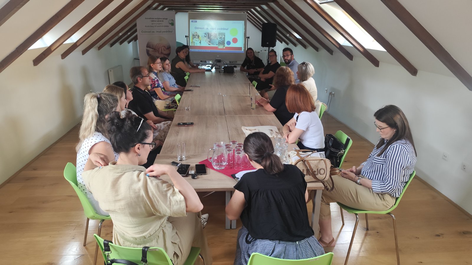 Na fotografiji je prikazan sastanak ili konferencija održana u potkrovnoj dvorani. Oko velikog drvenog stola sjedi petnaestak sudionika koji prate prezentaciju projiciranu na platnu na kraju prostorije. U pozadini se vidi i promotivni roll-up s nazivom projekta „Odmori se, imaš podršku“. Sudionici sjede u opuštenoj, radnoj atmosferi, a na stolu se nalaze čaše, vrčevi s vodom i osobni predmeti, što upućuje na radni sastanak ili završnu konferenciju projekta. Prostorija je osvijetljena prirodnim svjetlom koje dolazi kroz krovne prozore, stvarajući ugodnu i profesionalnu atmosferu.