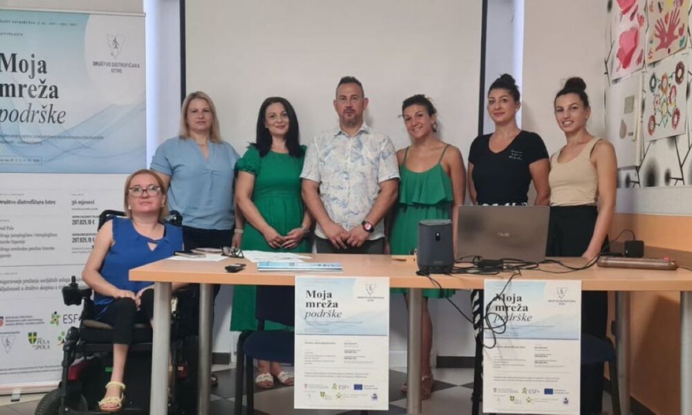 Na fotografiji je zabilježeno predstavljanje projekta „Moja mreža podrške“ u prostorijama Društva distrofičara Istre. Za stolom i ispred promotivnog panoa pozira sedam članova projektnog tima i suradnika, dok je jedna sudionica u prvom planu u invalidskim kolicima. Na stolu se nalaze prijenosno računalo, zvučnik i promotivni materijali projekta, a na prednjoj strani stola istaknuti su plakati s nazivom projekta. U pozadini se vidi projekcijsko platno te zid ukrašen dječjim likovnim radovima, što prostoru daje ugodnu i zajedničku atmosferu. Fotografija simbolizira početak provedbe projekta usmjerenog na pružanje podrške osobama s invaliditetom i njihovim obiteljima.