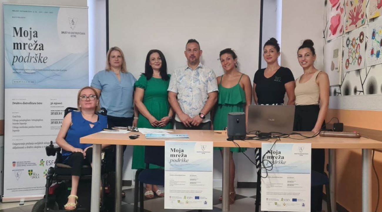 Na fotografiji je zabilježeno predstavljanje projekta „Moja mreža podrške“ u prostorijama Društva distrofičara Istre. Za stolom i ispred promotivnog panoa pozira sedam članova projektnog tima i suradnika, dok je jedna sudionica u prvom planu u invalidskim kolicima. Na stolu se nalaze prijenosno računalo, zvučnik i promotivni materijali projekta, a na prednjoj strani stola istaknuti su plakati s nazivom projekta. U pozadini se vidi projekcijsko platno te zid ukrašen dječjim likovnim radovima, što prostoru daje ugodnu i zajedničku atmosferu. Fotografija simbolizira početak provedbe projekta usmjerenog na pružanje podrške osobama s invaliditetom i njihovim obiteljima.
