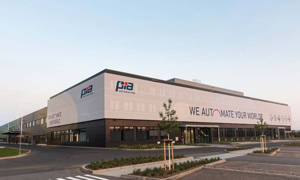 Na fotografiji se nalazi moderna poslovno-proizvodna zgrada tvrtke PIA Automation. Riječ je o velikom industrijskom objektu suvremenog dizajna, s bijelo-sivom fasadom i velikim logotipima tvrtke postavljenima na pročelju. Na zgradi se ističe slogan „WE AUTOMATE YOUR WORLD“, koji naglašava djelatnost tvrtke u području automatizacije. Ispred zgrade nalazi se uređeno parkiralište s asfaltiranim prometnicama, pješačkim prijelazima, mladim stablima i zelenim površinama s posađenim cvijećem. Cijeli prostor djeluje uredno, prostrano i moderno, ostavljajući dojam tehnološki naprednog i dobro organiziranog poslovnog centra. Fotografija je snimljena za vedrog i sunčanog dana, što dodatno ističe impresivnost i veličinu objekta.