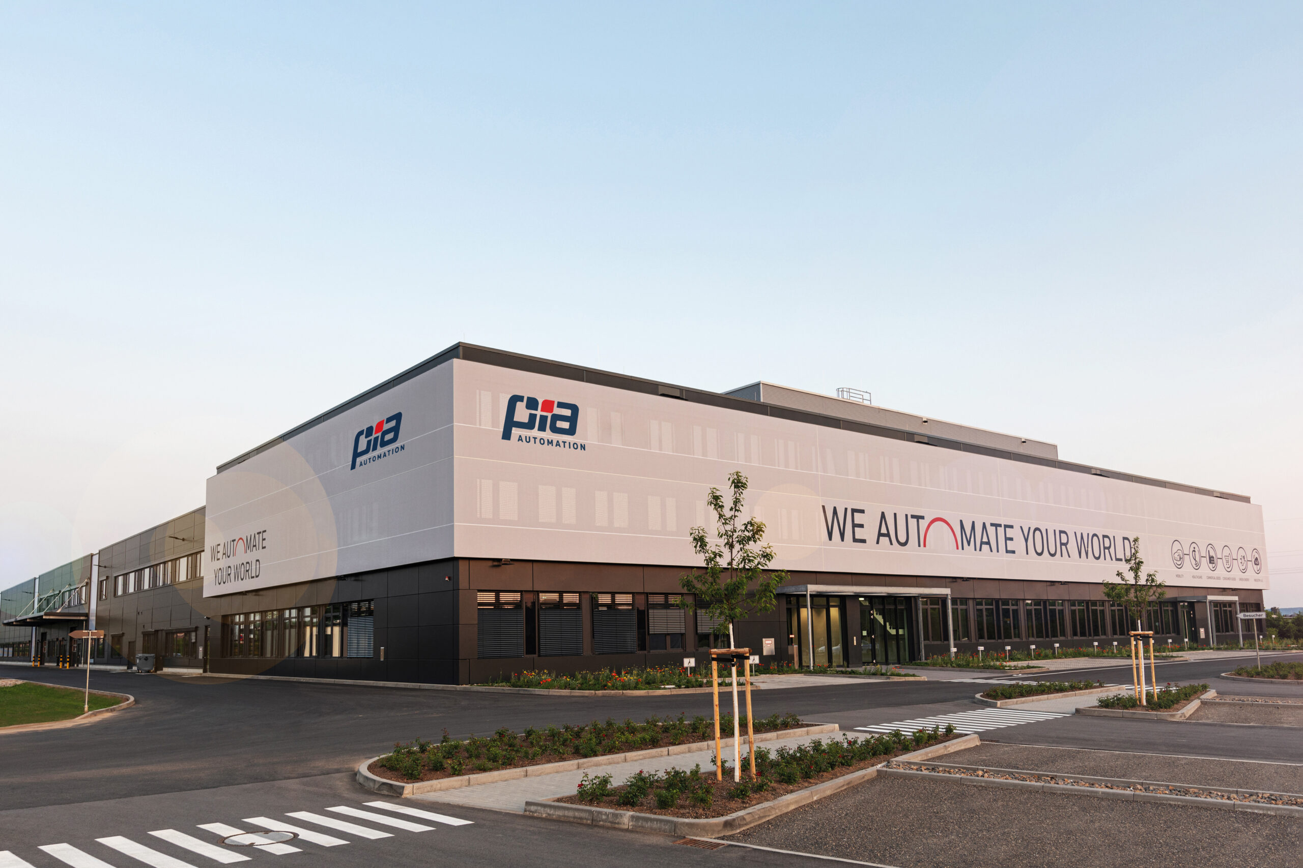 Na fotografiji se nalazi moderna poslovno-proizvodna zgrada tvrtke PIA Automation. Riječ je o velikom industrijskom objektu suvremenog dizajna, s bijelo-sivom fasadom i velikim logotipima tvrtke postavljenima na pročelju. Na zgradi se ističe slogan „WE AUTOMATE YOUR WORLD“, koji naglašava djelatnost tvrtke u području automatizacije. Ispred zgrade nalazi se uređeno parkiralište s asfaltiranim prometnicama, pješačkim prijelazima, mladim stablima i zelenim površinama s posađenim cvijećem. Cijeli prostor djeluje uredno, prostrano i moderno, ostavljajući dojam tehnološki naprednog i dobro organiziranog poslovnog centra. Fotografija je snimljena za vedrog i sunčanog dana, što dodatno ističe impresivnost i veličinu objekta.