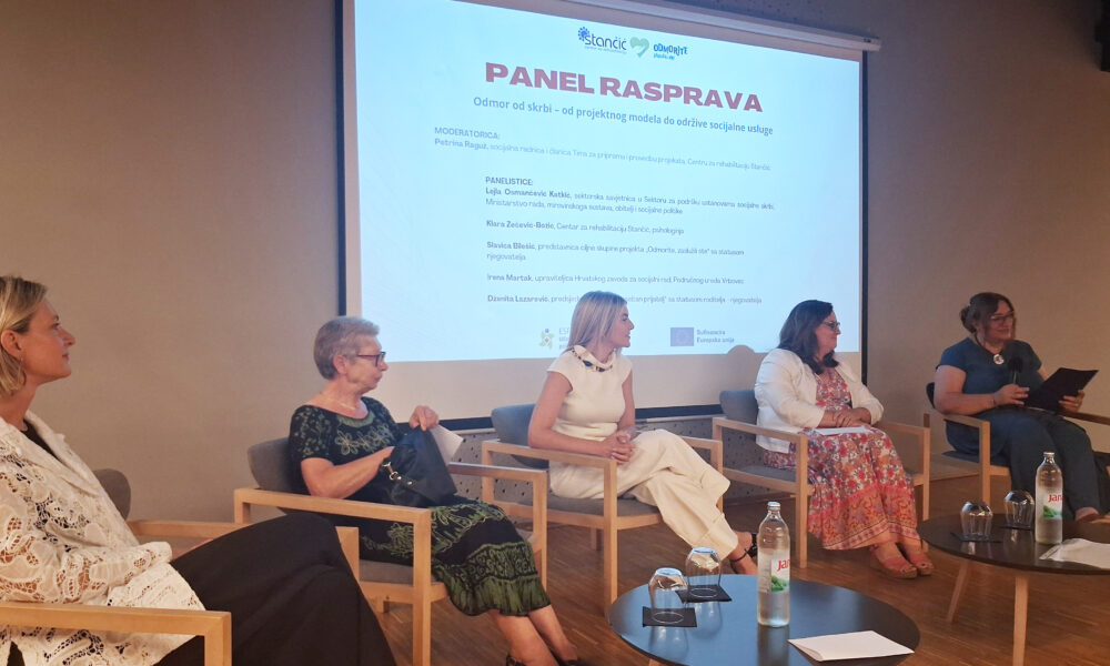 Sudionice panel rasprave sjede na pozornici ispred projekcije s naslovom „Odmor od skrbi – od projektnog modela do održive socijalne usluge”. Rasprava je posvećena razvoju usluge predaha za njegovatelje, a ispred sudionica nalaze se stolovi s vodom i čašama.