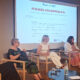 Sudionice panel rasprave sjede na pozornici ispred projekcije s naslovom „Odmor od skrbi – od projektnog modela do održive socijalne usluge”. Rasprava je posvećena razvoju usluge predaha za njegovatelje, a ispred sudionica nalaze se stolovi s vodom i čašama.