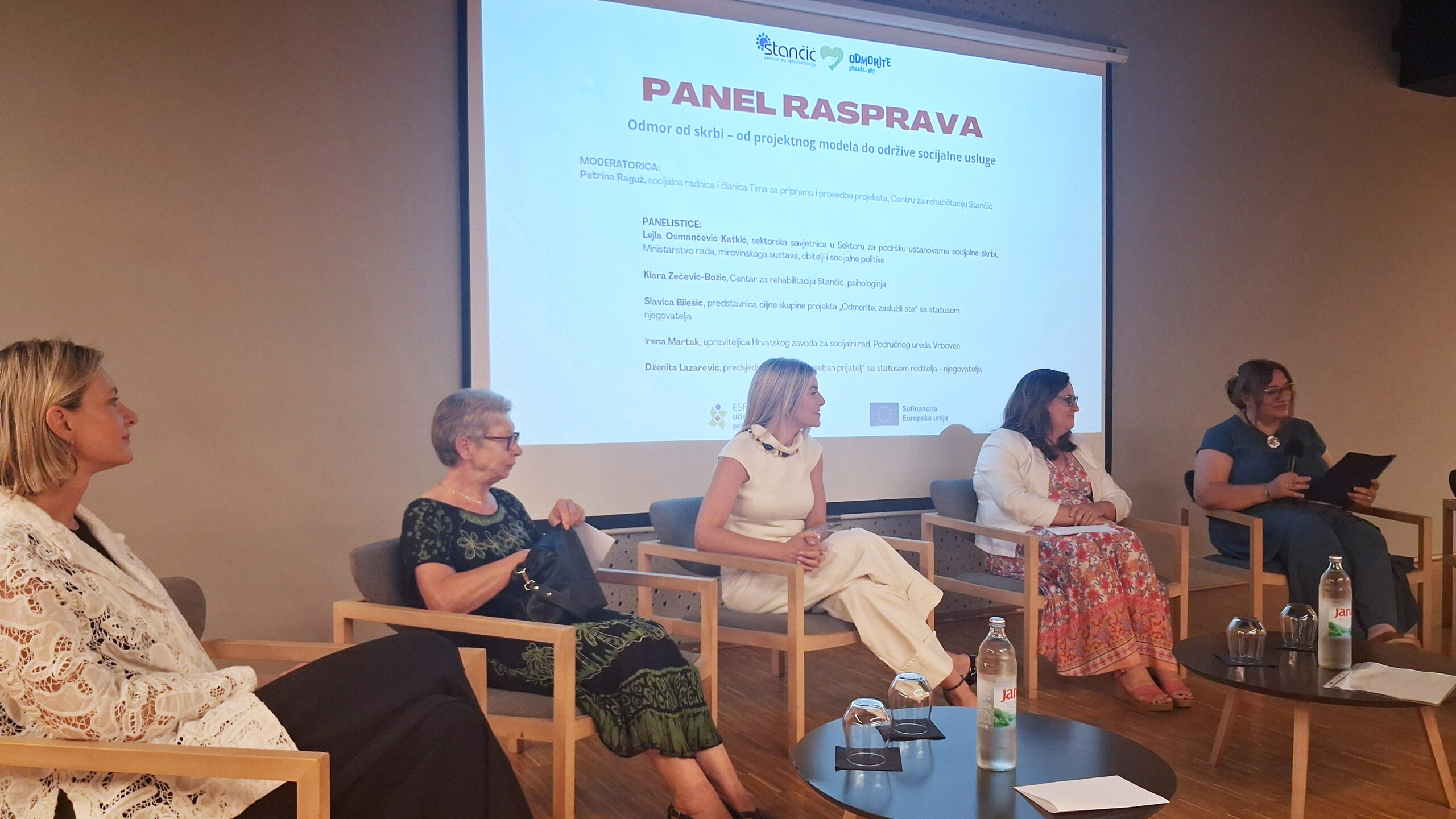 Sudionice panel rasprave sjede na pozornici ispred projekcije s naslovom „Odmor od skrbi – od projektnog modela do održive socijalne usluge”. Rasprava je posvećena razvoju usluge predaha za njegovatelje, a ispred sudionica nalaze se stolovi s vodom i čašama.