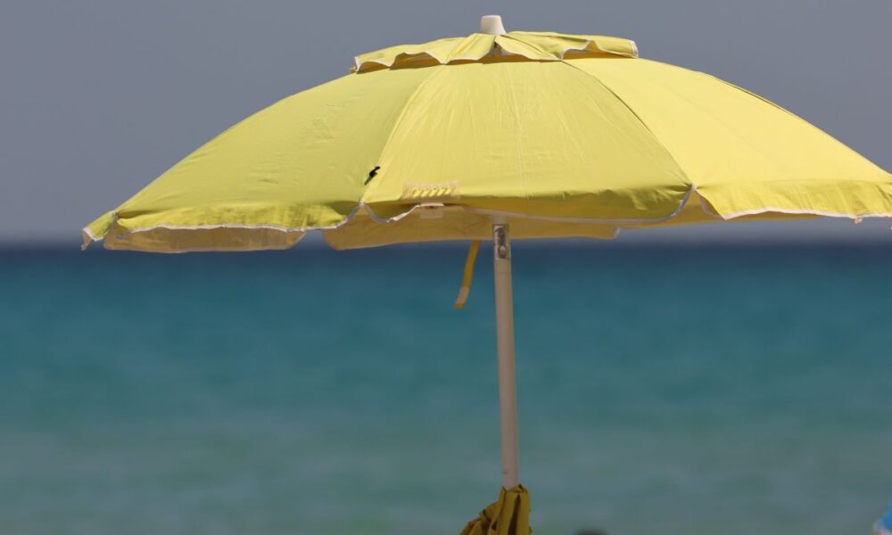 Na pješčanoj plaži stoji otvoreni žuti suncobran, ispod kojeg se nalaze torba, odjeća i par tenisica. U pozadini se vidi mirno more i vedro nebo.
