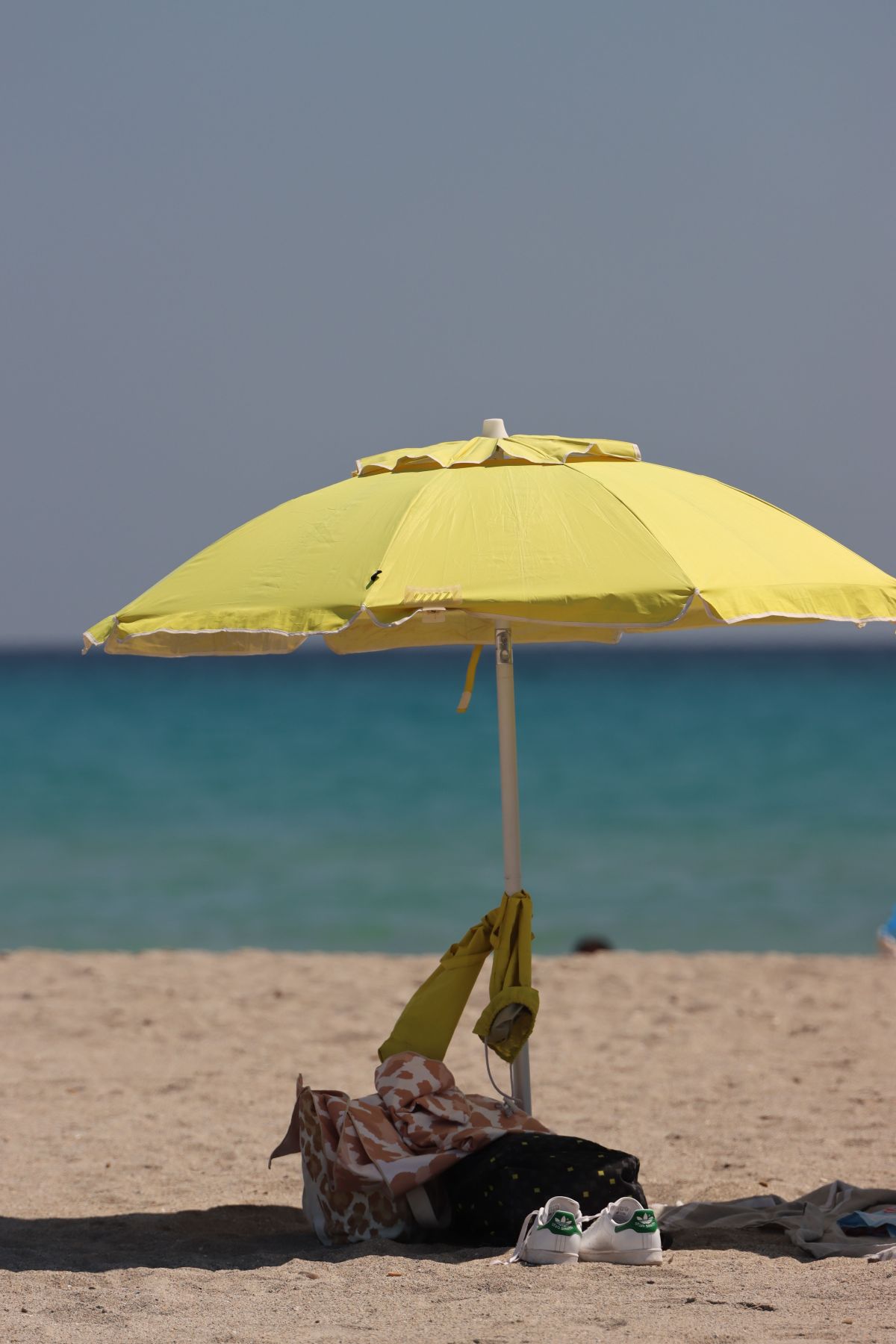Na pješčanoj plaži stoji otvoreni žuti suncobran, ispod kojeg se nalaze torba, odjeća i par tenisica. U pozadini se vidi mirno more i vedro nebo.