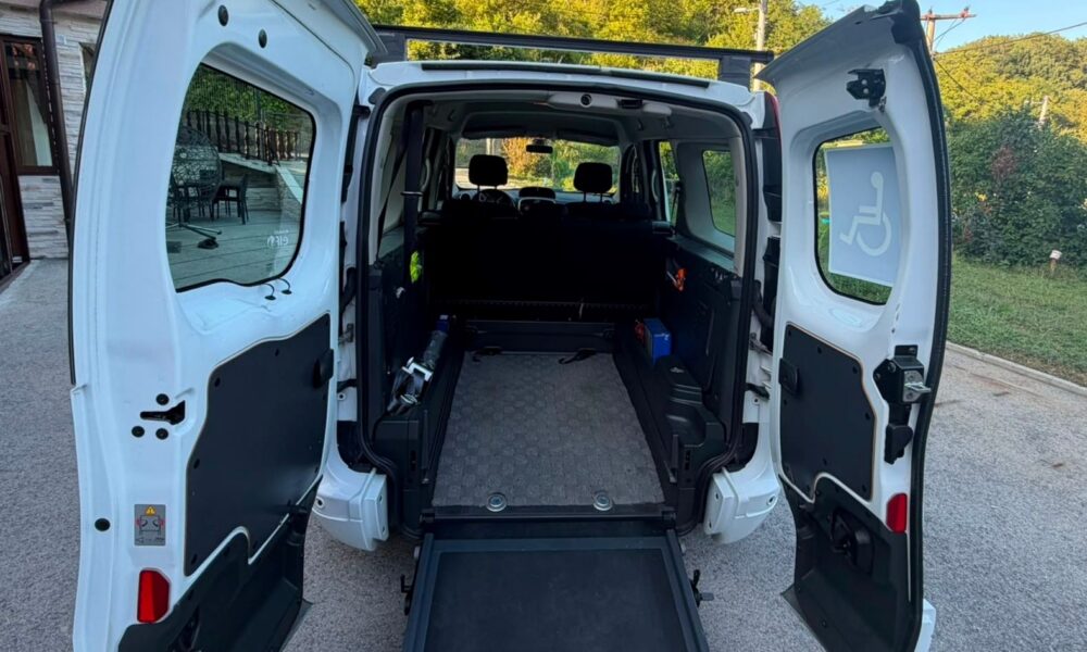 Na fotografiji je prikazan stražnji dio bijelog prilagođenog kombi-vozila s potpuno otvorenim dvokrilnim vratima i spuštenom rampom za ulazak osoba u invalidskim kolicima. U unutrašnjosti vozila vidi se prostran prostor s podnim vodilicama i sustavom za pričvršćivanje invalidskih kolica, što omogućuje siguran prijevoz osobe koja ostaje u kolicima tijekom vožnje. Iza prostora za kolica nalaze se sjedala za ostale putnike. Na desnom stražnjem prozoru istaknuta je međunarodno prepoznatljiva oznaka pristupačnosti za osobe s invaliditetom. Vozilo je parkirano ispred kuće, a u pozadini se vide zelenilo i šumoviti brežuljci pod vedrim plavim nebom. Fotografija jasno prikazuje prilagodbu vozila koja osobama s invaliditetom omogućuje lakši, sigurniji i dostojanstveniji ulazak u automobil te veću samostalnost i mobilnost.