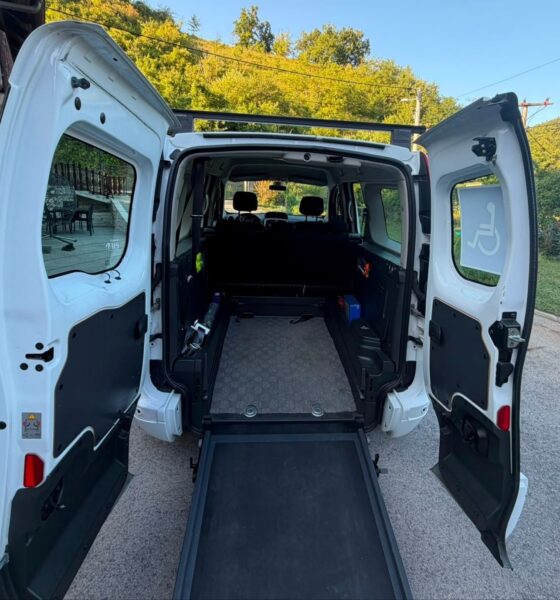 Na fotografiji je prikazan stražnji dio bijelog prilagođenog kombi-vozila s potpuno otvorenim dvokrilnim vratima i spuštenom rampom za ulazak osoba u invalidskim kolicima. U unutrašnjosti vozila vidi se prostran prostor s podnim vodilicama i sustavom za pričvršćivanje invalidskih kolica, što omogućuje siguran prijevoz osobe koja ostaje u kolicima tijekom vožnje. Iza prostora za kolica nalaze se sjedala za ostale putnike. Na desnom stražnjem prozoru istaknuta je međunarodno prepoznatljiva oznaka pristupačnosti za osobe s invaliditetom. Vozilo je parkirano ispred kuće, a u pozadini se vide zelenilo i šumoviti brežuljci pod vedrim plavim nebom. Fotografija jasno prikazuje prilagodbu vozila koja osobama s invaliditetom omogućuje lakši, sigurniji i dostojanstveniji ulazak u automobil te veću samostalnost i mobilnost.