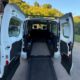 Na fotografiji je prikazan stražnji dio bijelog prilagođenog kombi-vozila s potpuno otvorenim dvokrilnim vratima i spuštenom rampom za ulazak osoba u invalidskim kolicima. U unutrašnjosti vozila vidi se prostran prostor s podnim vodilicama i sustavom za pričvršćivanje invalidskih kolica, što omogućuje siguran prijevoz osobe koja ostaje u kolicima tijekom vožnje. Iza prostora za kolica nalaze se sjedala za ostale putnike. Na desnom stražnjem prozoru istaknuta je međunarodno prepoznatljiva oznaka pristupačnosti za osobe s invaliditetom. Vozilo je parkirano ispred kuće, a u pozadini se vide zelenilo i šumoviti brežuljci pod vedrim plavim nebom. Fotografija jasno prikazuje prilagodbu vozila koja osobama s invaliditetom omogućuje lakši, sigurniji i dostojanstveniji ulazak u automobil te veću samostalnost i mobilnost.
