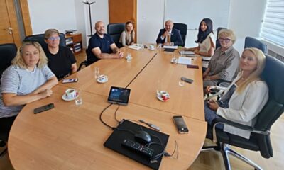 Na fotografiji se nalazi osam osoba okupljenih oko velikog konferencijskog stola u prostoriji za sastanke. Sudionici sjede s obje strane stola, a ispred njih se nalaze šalice kave, čaše s vodom i mobilni telefoni. U središtu stola nalazi se konferencijski uređaj za komunikaciju. U pozadini se vide uredski elementi poput ormara, vješalice i prozora s roletama, što upućuje na to da je fotografija snimljena tijekom službenog radnog sastanka. Sudionici su opušteni i gledaju prema kameri, ostavljajući dojam konstruktivnog i radnog ozračja tijekom razgovora o važnim temama vezanim uz osobe s invaliditetom.