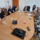 Na fotografiji se nalazi osam osoba okupljenih oko velikog konferencijskog stola u prostoriji za sastanke. Sudionici sjede s obje strane stola, a ispred njih se nalaze šalice kave, čaše s vodom i mobilni telefoni. U središtu stola nalazi se konferencijski uređaj za komunikaciju. U pozadini se vide uredski elementi poput ormara, vješalice i prozora s roletama, što upućuje na to da je fotografija snimljena tijekom službenog radnog sastanka. Sudionici su opušteni i gledaju prema kameri, ostavljajući dojam konstruktivnog i radnog ozračja tijekom razgovora o važnim temama vezanim uz osobe s invaliditetom.