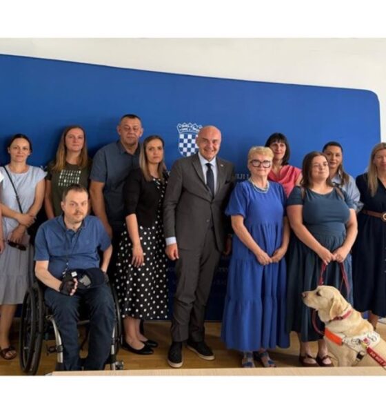 Na fotografiji je skupina ljudi okupljena u službenom prostoru ispred plave pozadine s grbom Republike Hrvatske. U središtu fotografije nalazi se muškarac u tamnom odijelu, okružen predstavnicima udruga i sudionicima sastanka. Među okupljenima su osobe različite životne dobi, uključujući jednu osobu u invalidskim kolicima te psa vodiča koji sjedi u prvom planu uz jednu od sudionica. Svi su okrenuti prema kameri i poziraju za zajedničku fotografiju nakon sastanka. Atmosfera je formalna, ali srdačna, što upućuje na službeni susret posvećen temama od interesa za osobe s invaliditetom.