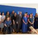 Na fotografiji je skupina ljudi okupljena u službenom prostoru ispred plave pozadine s grbom Republike Hrvatske. U središtu fotografije nalazi se muškarac u tamnom odijelu, okružen predstavnicima udruga i sudionicima sastanka. Među okupljenima su osobe različite životne dobi, uključujući jednu osobu u invalidskim kolicima te psa vodiča koji sjedi u prvom planu uz jednu od sudionica. Svi su okrenuti prema kameri i poziraju za zajedničku fotografiju nakon sastanka. Atmosfera je formalna, ali srdačna, što upućuje na službeni susret posvećen temama od interesa za osobe s invaliditetom.