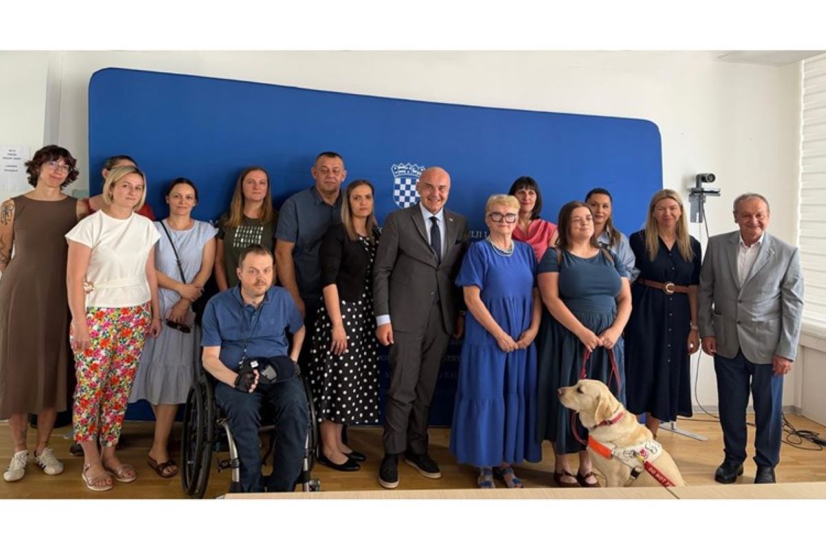 Na fotografiji je skupina ljudi okupljena u službenom prostoru ispred plave pozadine s grbom Republike Hrvatske. U središtu fotografije nalazi se muškarac u tamnom odijelu, okružen predstavnicima udruga i sudionicima sastanka. Među okupljenima su osobe različite životne dobi, uključujući jednu osobu u invalidskim kolicima te psa vodiča koji sjedi u prvom planu uz jednu od sudionica. Svi su okrenuti prema kameri i poziraju za zajedničku fotografiju nakon sastanka. Atmosfera je formalna, ali srdačna, što upućuje na službeni susret posvećen temama od interesa za osobe s invaliditetom.