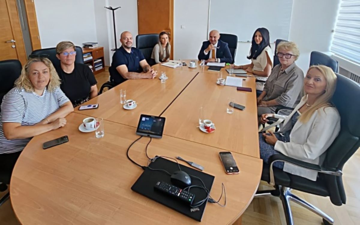 Na fotografiji se nalazi osam osoba okupljenih oko velikog konferencijskog stola u prostoriji za sastanke. Sudionici sjede s obje strane stola, a ispred njih se nalaze šalice kave, čaše s vodom i mobilni telefoni. U središtu stola nalazi se konferencijski uređaj za komunikaciju. U pozadini se vide uredski elementi poput ormara, vješalice i prozora s roletama, što upućuje na to da je fotografija snimljena tijekom službenog radnog sastanka. Sudionici su opušteni i gledaju prema kameri, ostavljajući dojam konstruktivnog i radnog ozračja tijekom razgovora o važnim temama vezanim uz osobe s invaliditetom.