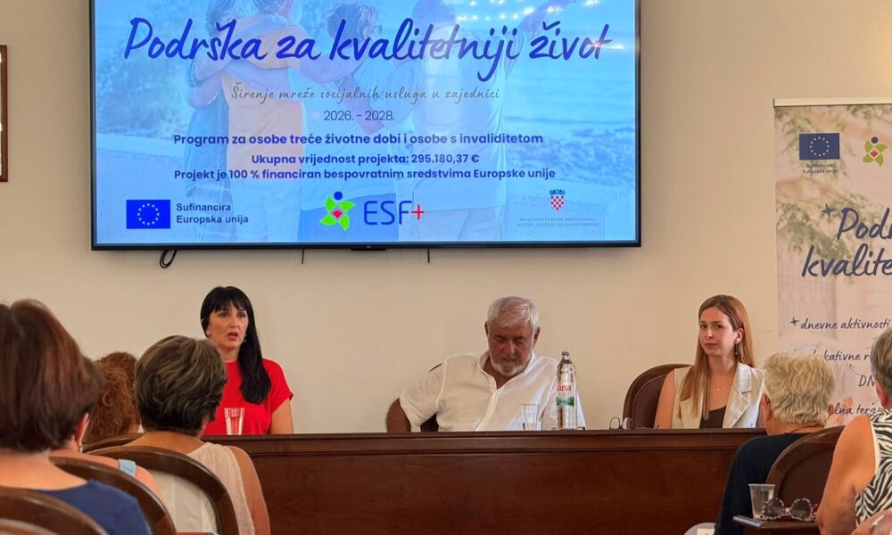 Na fotografiji je prikazan trenutak s početne konferencije projekta „Podrška za kvalitetniji život“. U prvom planu za govornicom sjede tri osobe koje predstavljaju projekt okupljenim sudionicima. Lijevo govornica izlaže projekt, dok ostali članovi panela pažljivo prate izlaganje. Iznad njih nalazi se veliki ekran na kojem je prikazana naslovna prezentacija projekta s nazivom „Podrška za kvalitetniji život“, logotipima Udruge osoba s invaliditetom TOMS, Grada Trogira, ESF+ programa i Europske unije te osnovnim informacijama o projektu. U dvorani se vidi publika koja prati konferenciju, a s desne strane nalazi se promotivni roll-up projekta. Fotografija prikazuje službenu i radnu atmosferu predstavljanja projekta, uz velik interes okupljenih sudionika.
