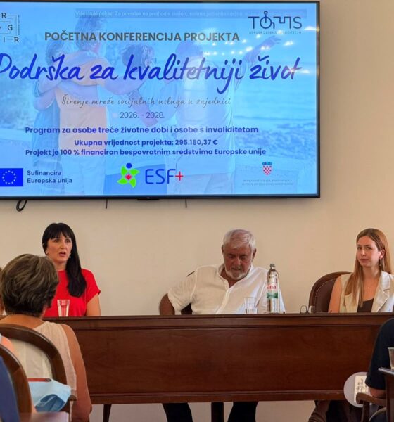 Na fotografiji je prikazan trenutak s početne konferencije projekta „Podrška za kvalitetniji život“. U prvom planu za govornicom sjede tri osobe koje predstavljaju projekt okupljenim sudionicima. Lijevo govornica izlaže projekt, dok ostali članovi panela pažljivo prate izlaganje. Iznad njih nalazi se veliki ekran na kojem je prikazana naslovna prezentacija projekta s nazivom „Podrška za kvalitetniji život“, logotipima Udruge osoba s invaliditetom TOMS, Grada Trogira, ESF+ programa i Europske unije te osnovnim informacijama o projektu. U dvorani se vidi publika koja prati konferenciju, a s desne strane nalazi se promotivni roll-up projekta. Fotografija prikazuje službenu i radnu atmosferu predstavljanja projekta, uz velik interes okupljenih sudionika.