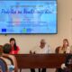 Na fotografiji je prikazan trenutak s početne konferencije projekta „Podrška za kvalitetniji život“. U prvom planu za govornicom sjede tri osobe koje predstavljaju projekt okupljenim sudionicima. Lijevo govornica izlaže projekt, dok ostali članovi panela pažljivo prate izlaganje. Iznad njih nalazi se veliki ekran na kojem je prikazana naslovna prezentacija projekta s nazivom „Podrška za kvalitetniji život“, logotipima Udruge osoba s invaliditetom TOMS, Grada Trogira, ESF+ programa i Europske unije te osnovnim informacijama o projektu. U dvorani se vidi publika koja prati konferenciju, a s desne strane nalazi se promotivni roll-up projekta. Fotografija prikazuje službenu i radnu atmosferu predstavljanja projekta, uz velik interes okupljenih sudionika.