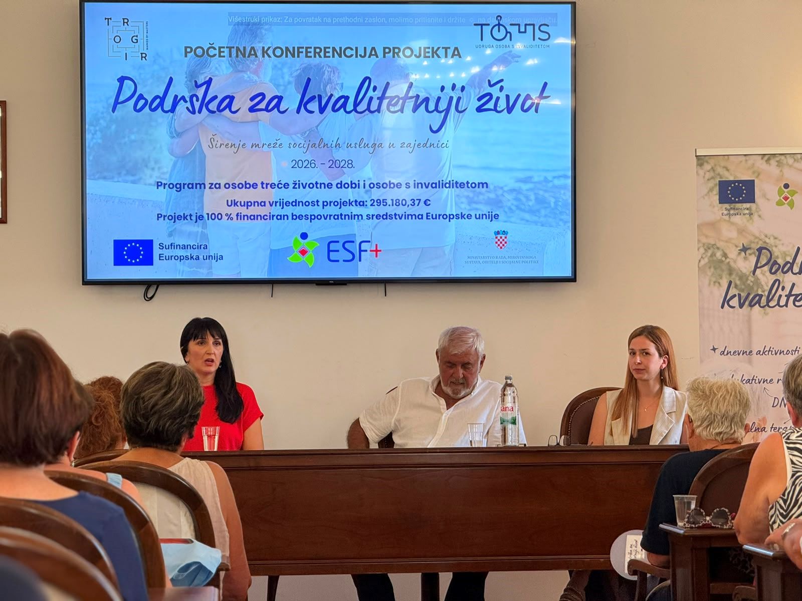 Na fotografiji je prikazan trenutak s početne konferencije projekta „Podrška za kvalitetniji život“. U prvom planu za govornicom sjede tri osobe koje predstavljaju projekt okupljenim sudionicima. Lijevo govornica izlaže projekt, dok ostali članovi panela pažljivo prate izlaganje. Iznad njih nalazi se veliki ekran na kojem je prikazana naslovna prezentacija projekta s nazivom „Podrška za kvalitetniji život“, logotipima Udruge osoba s invaliditetom TOMS, Grada Trogira, ESF+ programa i Europske unije te osnovnim informacijama o projektu. U dvorani se vidi publika koja prati konferenciju, a s desne strane nalazi se promotivni roll-up projekta. Fotografija prikazuje službenu i radnu atmosferu predstavljanja projekta, uz velik interes okupljenih sudionika.