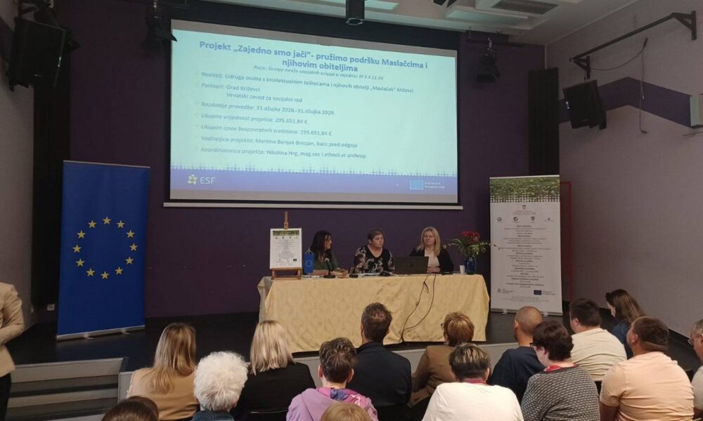 Na fotografiji je prikazano predstavljanje projekta u konferencijskoj dvorani pred okupljenim sudionicima. U središtu pozornice za stolom sjede tri žene koje sudjeluju u predstavljanju projekta. Iza njih se na velikom projekcijskom platnu prikazuje prezentacija s naslovom „Projekt 'Zajedno smo jači' – pružimo podršku Maslačcima i njihovim obiteljima“. Na slajdu su vidljive osnovne informacije o projektu, uključujući nositelja projekta, partnere, razdoblje provedbe, ukupnu vrijednost i voditeljice projekta. S lijeve strane pozornice nalazi se zastava Europske unije, dok je s desne strane postavljen promotivni roll-up s informacijama o projektu. Ispred stola nalazi se i uokvireni plakat projekta te mala zastavica Europske unije. U prvom planu vidi se publika koja pozorno prati predstavljanje, a na lijevoj strani fotografije nalazi se osoba koja fotografira ili snima događaj. Fotografija prikazuje službeno predstavljanje projekta financiranog sredstvima Europske unije, uz sudjelovanje predstavnika nositelja i partnera projekta te zainteresirane javnosti.