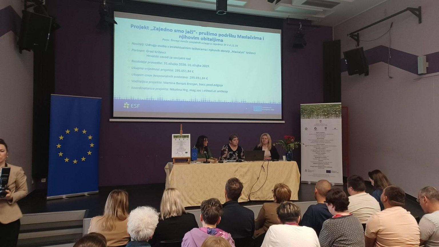 Na fotografiji je prikazano predstavljanje projekta u konferencijskoj dvorani pred okupljenim sudionicima. U središtu pozornice za stolom sjede tri žene koje sudjeluju u predstavljanju projekta. Iza njih se na velikom projekcijskom platnu prikazuje prezentacija s naslovom „Projekt 'Zajedno smo jači' – pružimo podršku Maslačcima i njihovim obiteljima“. Na slajdu su vidljive osnovne informacije o projektu, uključujući nositelja projekta, partnere, razdoblje provedbe, ukupnu vrijednost i voditeljice projekta. S lijeve strane pozornice nalazi se zastava Europske unije, dok je s desne strane postavljen promotivni roll-up s informacijama o projektu. Ispred stola nalazi se i uokvireni plakat projekta te mala zastavica Europske unije. U prvom planu vidi se publika koja pozorno prati predstavljanje, a na lijevoj strani fotografije nalazi se osoba koja fotografira ili snima događaj. Fotografija prikazuje službeno predstavljanje projekta financiranog sredstvima Europske unije, uz sudjelovanje predstavnika nositelja i partnera projekta te zainteresirane javnosti.