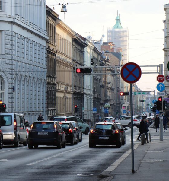 Na fotografiji je prikazana prometna ulica u središtu Zagreba s gustim prometom osobnih automobila i kombija. Uz kolnik se nalaze nogostupi kojima prolaze pješaci, dok se iznad ceste vide tramvajske žice karakteristične za zagrebački centar. Na raskrižju su vidljivi prometni znakovi i semafori s upaljenim crvenim svjetlom, što ukazuje na zaustavljen ili usporen promet. U pozadini se nazire toranj zagrebačke katedrale, djelomično prekriven zaštitnom skelom, što upućuje na obnovu nakon potresa. Fotografija prikazuje svakodnevni gradski život i prometnu dinamiku u povijesnoj jezgri Zagreba, s naglaskom na urbanu infrastrukturu, sudionike u prometu i arhitekturu koja okružuje ulicu.