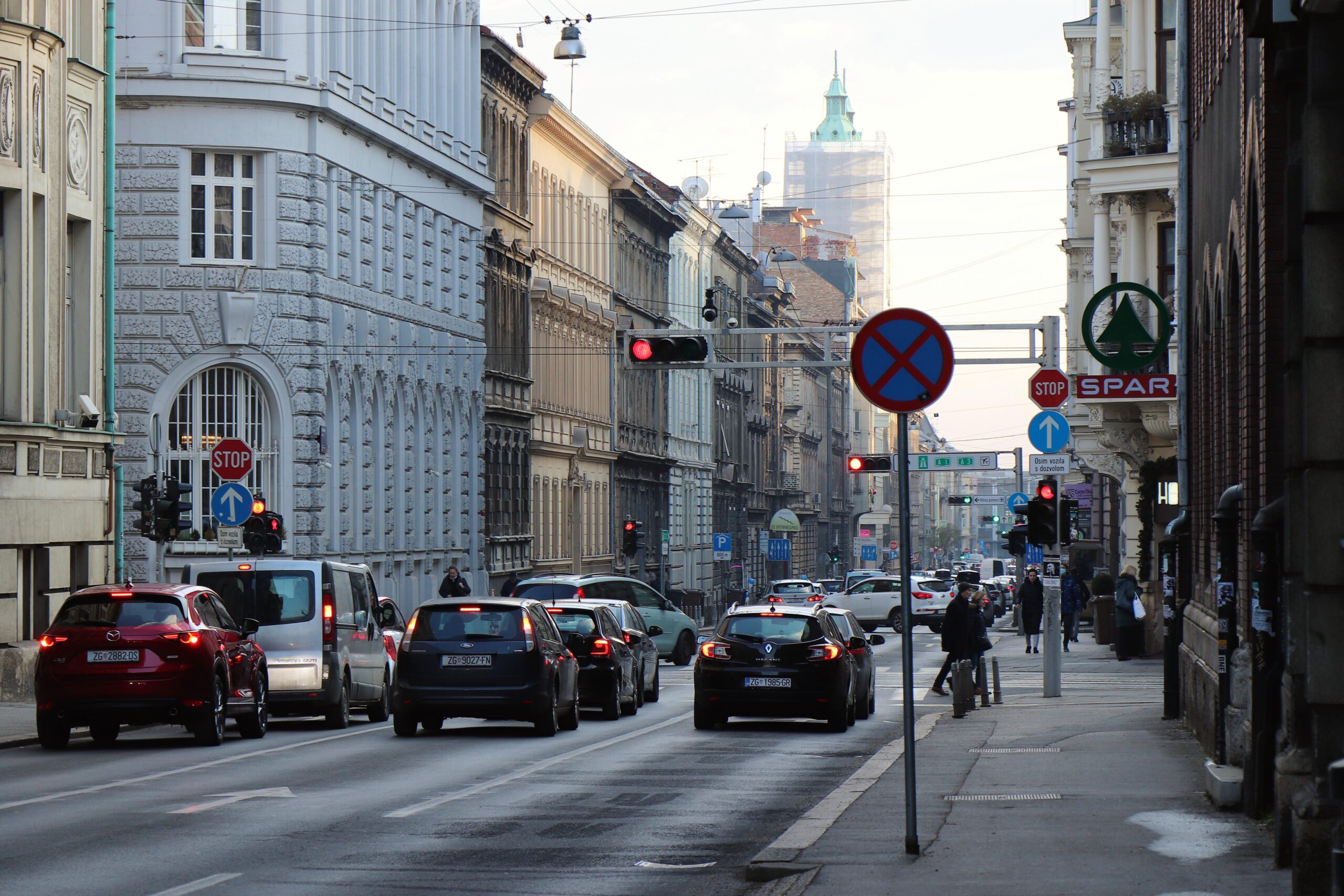 Na fotografiji je prikazana prometna ulica u središtu Zagreba s gustim prometom osobnih automobila i kombija. Uz kolnik se nalaze nogostupi kojima prolaze pješaci, dok se iznad ceste vide tramvajske žice karakteristične za zagrebački centar. Na raskrižju su vidljivi prometni znakovi i semafori s upaljenim crvenim svjetlom, što ukazuje na zaustavljen ili usporen promet. U pozadini se nazire toranj zagrebačke katedrale, djelomično prekriven zaštitnom skelom, što upućuje na obnovu nakon potresa. Fotografija prikazuje svakodnevni gradski život i prometnu dinamiku u povijesnoj jezgri Zagreba, s naglaskom na urbanu infrastrukturu, sudionike u prometu i arhitekturu koja okružuje ulicu.