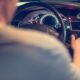 Na fotografiji se vidi unutrašnjost automobila iz perspektive stražnjeg sjedala. U prvom planu nalazi se vozač, čije su lice i tijelo zamućeni, dok desnom rukom poseže prema kontakt-bravi, vjerojatno kako bi upalio ili ugasio vozilo. U pozadini se jasno vide upravljač, osvijetljena instrumentna ploča s brzinomjerom i kontrolna ploča automobila. Na vjetrobranskom staklu postavljen je držač za mobitel, što sugerira da je riječ o taksi-vozilu ili automobilu koji se koristi za navigaciju. Fotografija stvara dojam svakodnevne vožnje i fokusira se na trenutak kada vozač upravlja ili priprema vozilo za polazak.