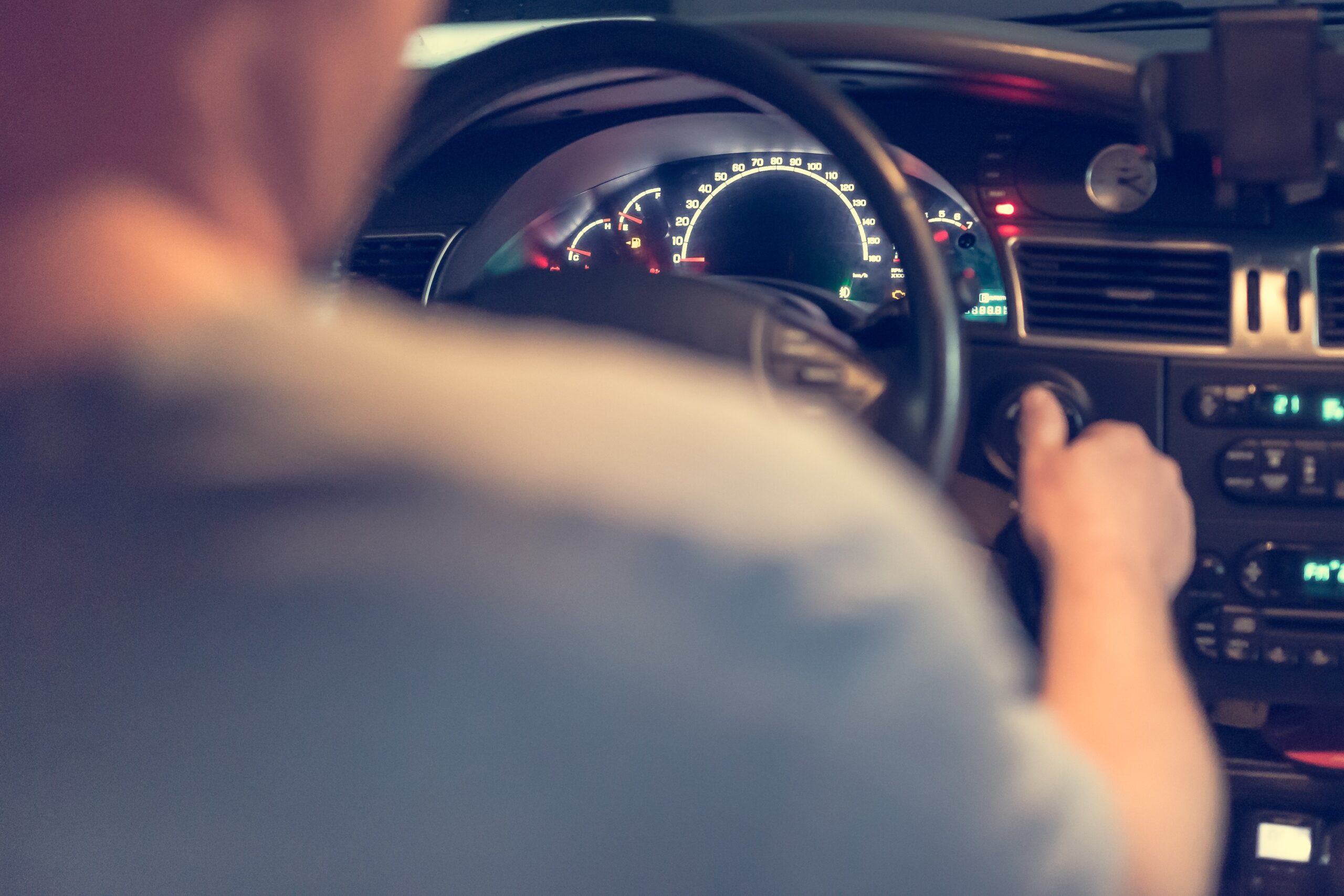 Na fotografiji se vidi unutrašnjost automobila iz perspektive stražnjeg sjedala. U prvom planu nalazi se vozač, čije su lice i tijelo zamućeni, dok desnom rukom poseže prema kontakt-bravi, vjerojatno kako bi upalio ili ugasio vozilo. U pozadini se jasno vide upravljač, osvijetljena instrumentna ploča s brzinomjerom i kontrolna ploča automobila. Na vjetrobranskom staklu postavljen je držač za mobitel, što sugerira da je riječ o taksi-vozilu ili automobilu koji se koristi za navigaciju. Fotografija stvara dojam svakodnevne vožnje i fokusira se na trenutak kada vozač upravlja ili priprema vozilo za polazak.
