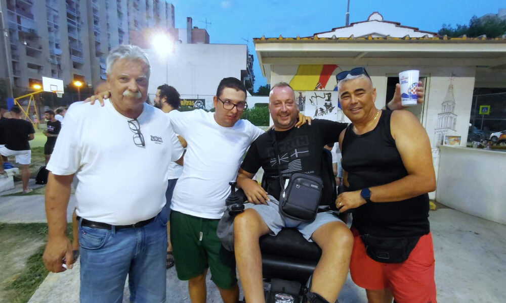 Četiri muškarca poziraju zagrljeni tijekom večernjeg druženja na sportskom igralištu. Jedan od njih sjedi u motornim invalidskim kolicima, dok se u pozadini vide košarkaški teren, okupljeni ljudi i okolne zgrade.