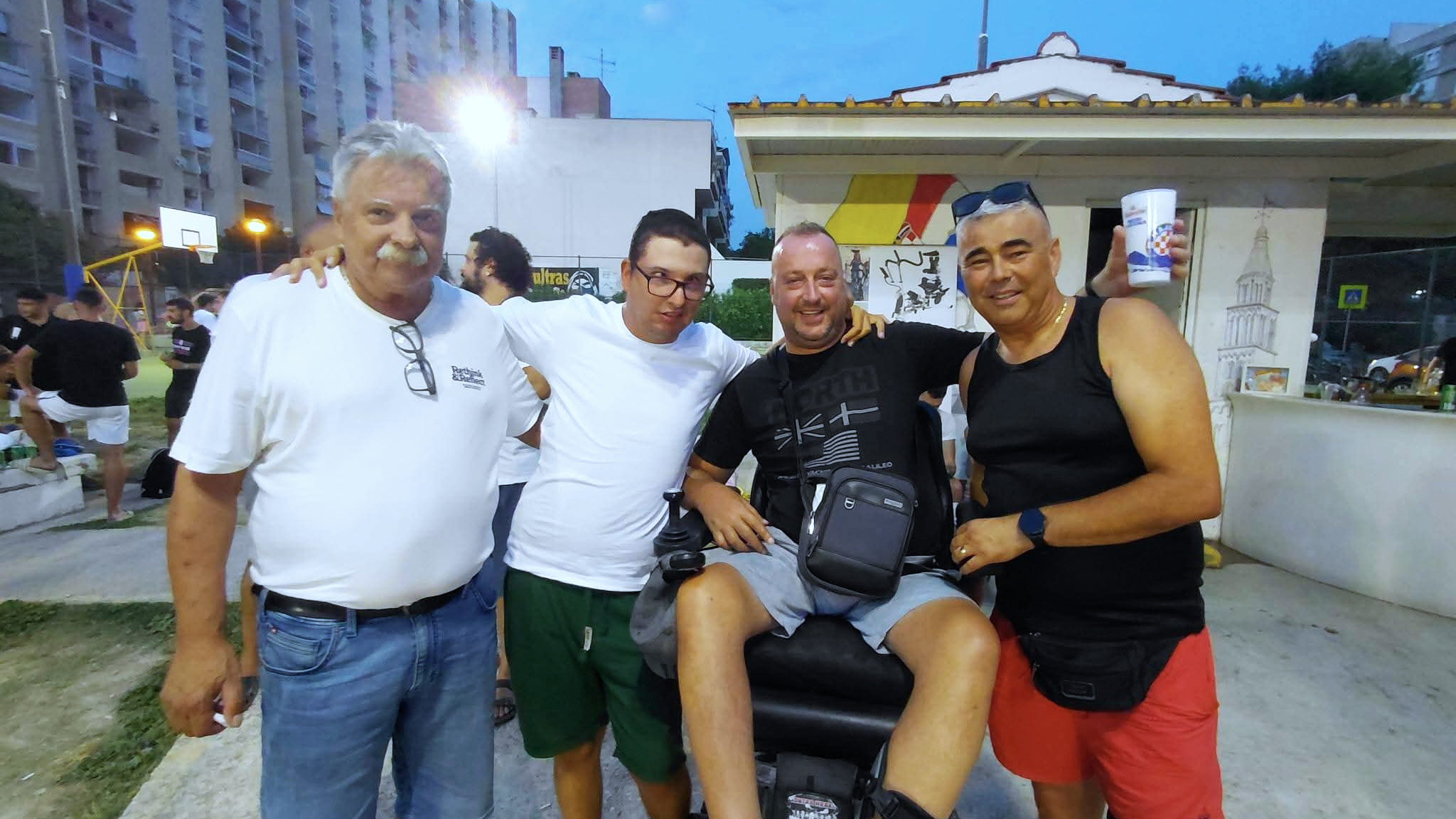 Četiri muškarca poziraju zagrljeni tijekom večernjeg druženja na sportskom igralištu. Jedan od njih sjedi u motornim invalidskim kolicima, dok se u pozadini vide košarkaški teren, okupljeni ljudi i okolne zgrade.