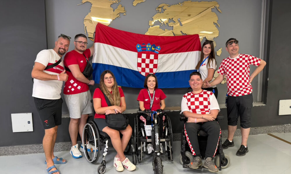 Članovi hrvatske boccia reprezentacije i stručnog stožera poziraju ispred hrvatske zastave. Dio sportaša nalazi se u sportskim kolicima, a svi su odjeveni u odjeću s hrvatskim nacionalnim obilježjima.