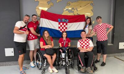 Članovi hrvatske boccia reprezentacije i stručnog stožera poziraju ispred hrvatske zastave. Dio sportaša nalazi se u sportskim kolicima, a svi su odjeveni u odjeću s hrvatskim nacionalnim obilježjima.