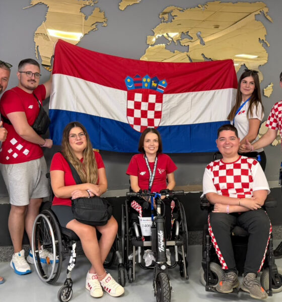 Članovi hrvatske boccia reprezentacije i stručnog stožera poziraju ispred hrvatske zastave. Dio sportaša nalazi se u sportskim kolicima, a svi su odjeveni u odjeću s hrvatskim nacionalnim obilježjima.