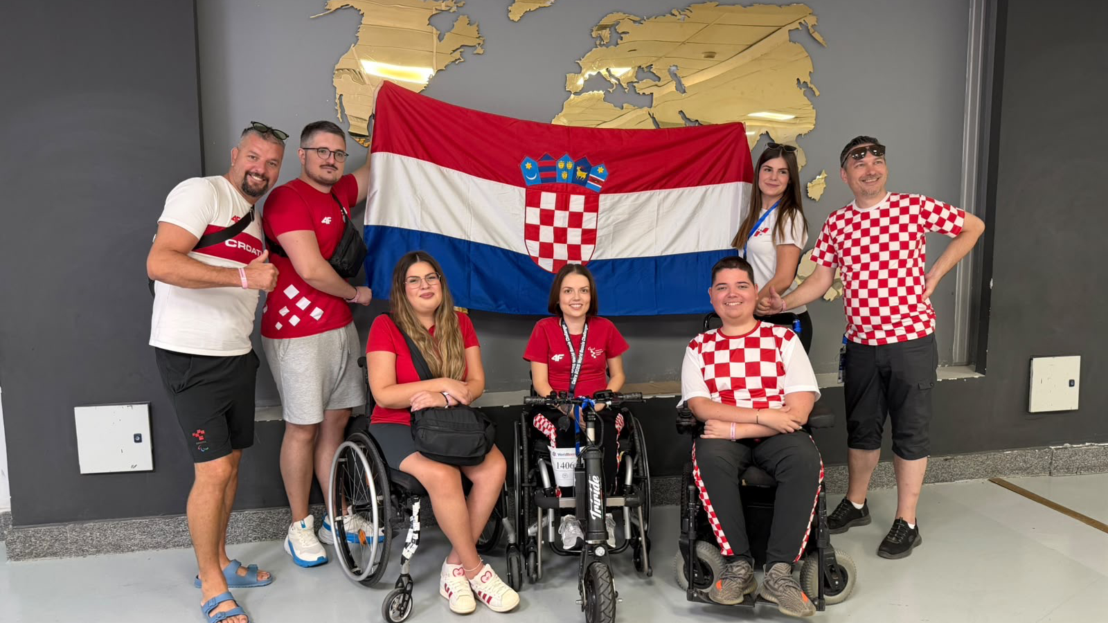 Članovi hrvatske boccia reprezentacije i stručnog stožera poziraju ispred hrvatske zastave. Dio sportaša nalazi se u sportskim kolicima, a svi su odjeveni u odjeću s hrvatskim nacionalnim obilježjima.