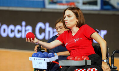 Hrvatska boccia reprezentativka u kolicima koncentrirano izvodi bacanje crvene lopte tijekom natjecanja. U pozadini se vidi protivnička igračica s plavim loptama te sportska dvorana u kojoj se održava natjecanje.