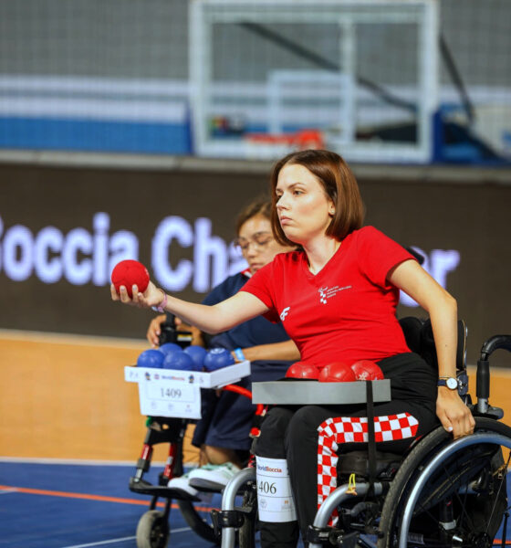 Hrvatska boccia reprezentativka u kolicima koncentrirano izvodi bacanje crvene lopte tijekom natjecanja. U pozadini se vidi protivnička igračica s plavim loptama te sportska dvorana u kojoj se održava natjecanje.