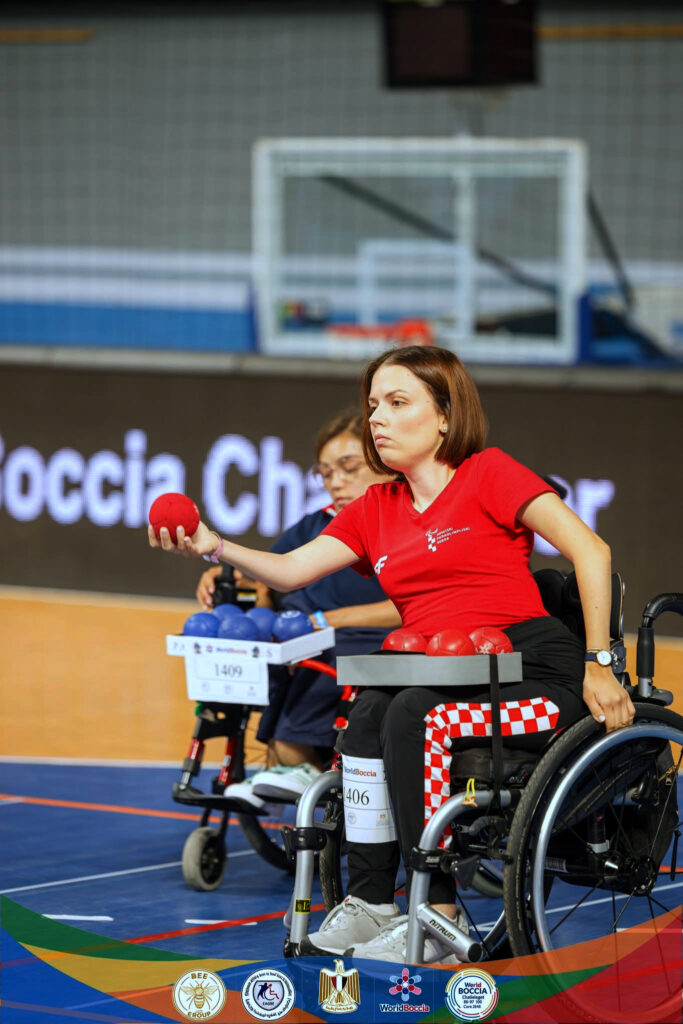 Hrvatska boccia reprezentativka u kolicima koncentrirano izvodi bacanje crvene lopte tijekom natjecanja. U pozadini se vidi protivnička igračica s plavim loptama te sportska dvorana u kojoj se održava natjecanje.