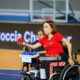 Hrvatska boccia reprezentativka u kolicima koncentrirano izvodi bacanje crvene lopte tijekom natjecanja. U pozadini se vidi protivnička igračica s plavim loptama te sportska dvorana u kojoj se održava natjecanje.