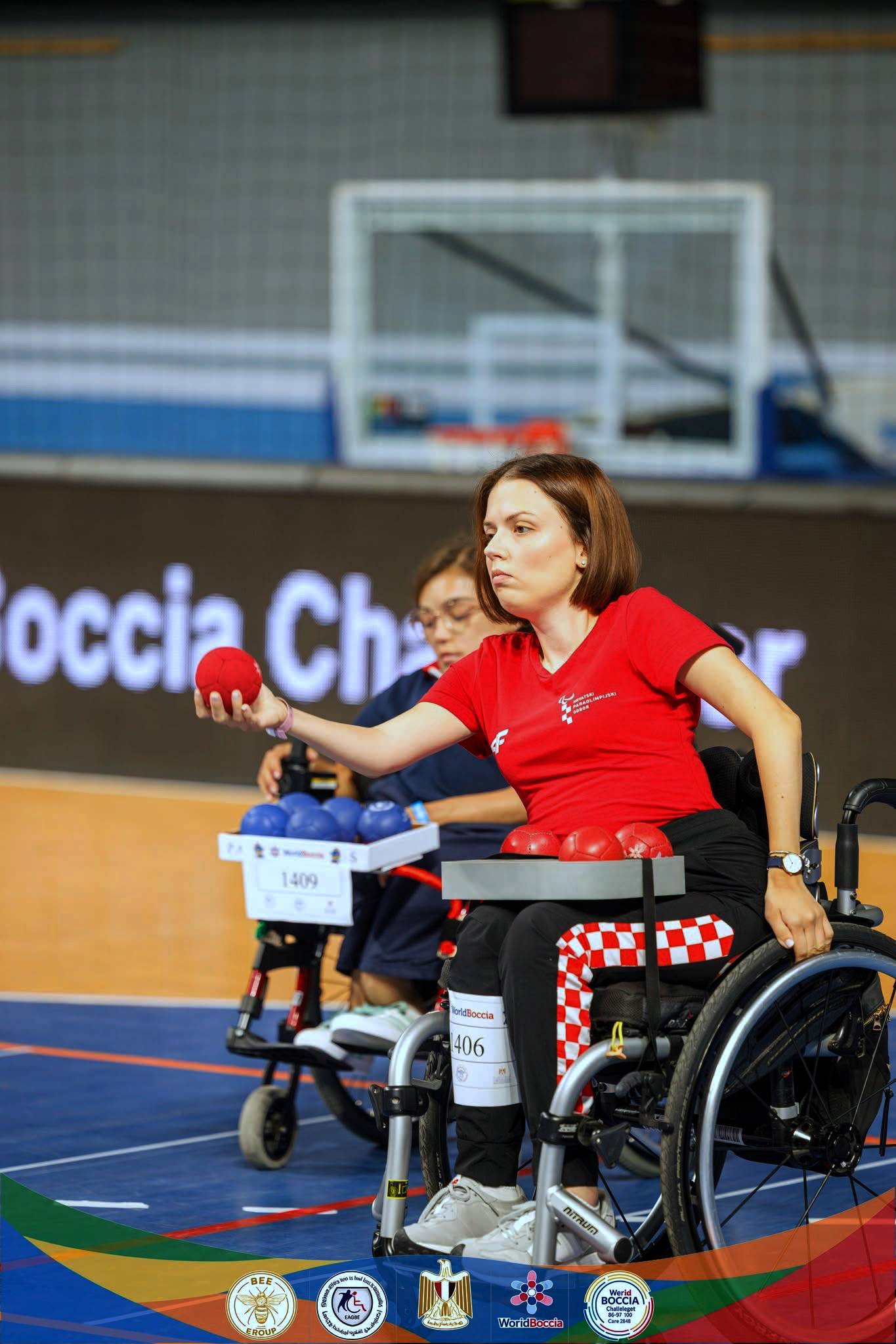 Hrvatska boccia reprezentativka u kolicima koncentrirano izvodi bacanje crvene lopte tijekom natjecanja. U pozadini se vidi protivnička igračica s plavim loptama te sportska dvorana u kojoj se održava natjecanje.