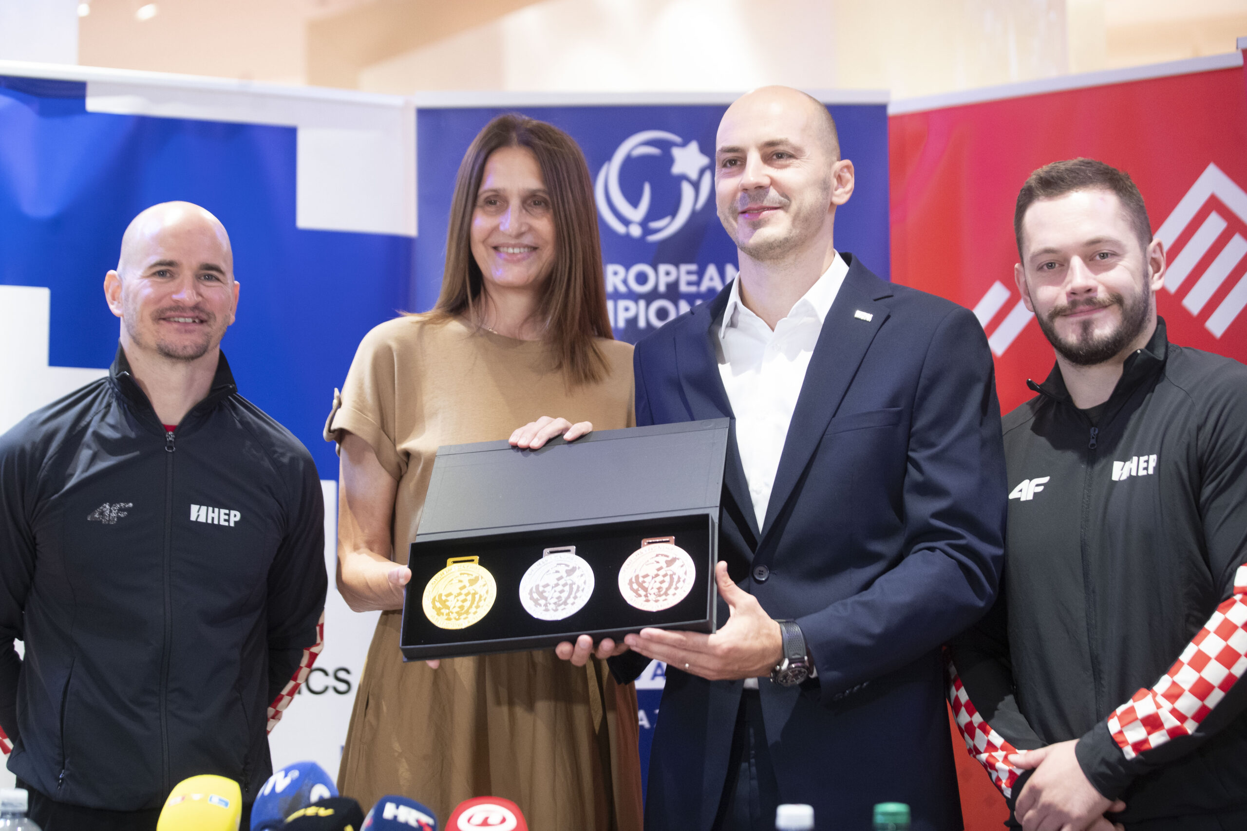 Četvero ljudi pozira na konferenciji za medije držeći kutiju sa zlatnom, srebrnom i brončanom medaljom.