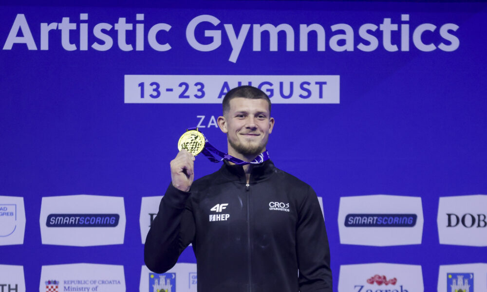 Gimnastičar pozira sa zlatnom medaljom ispred pozadine s natpisom „Artistic Gymnastics“.
