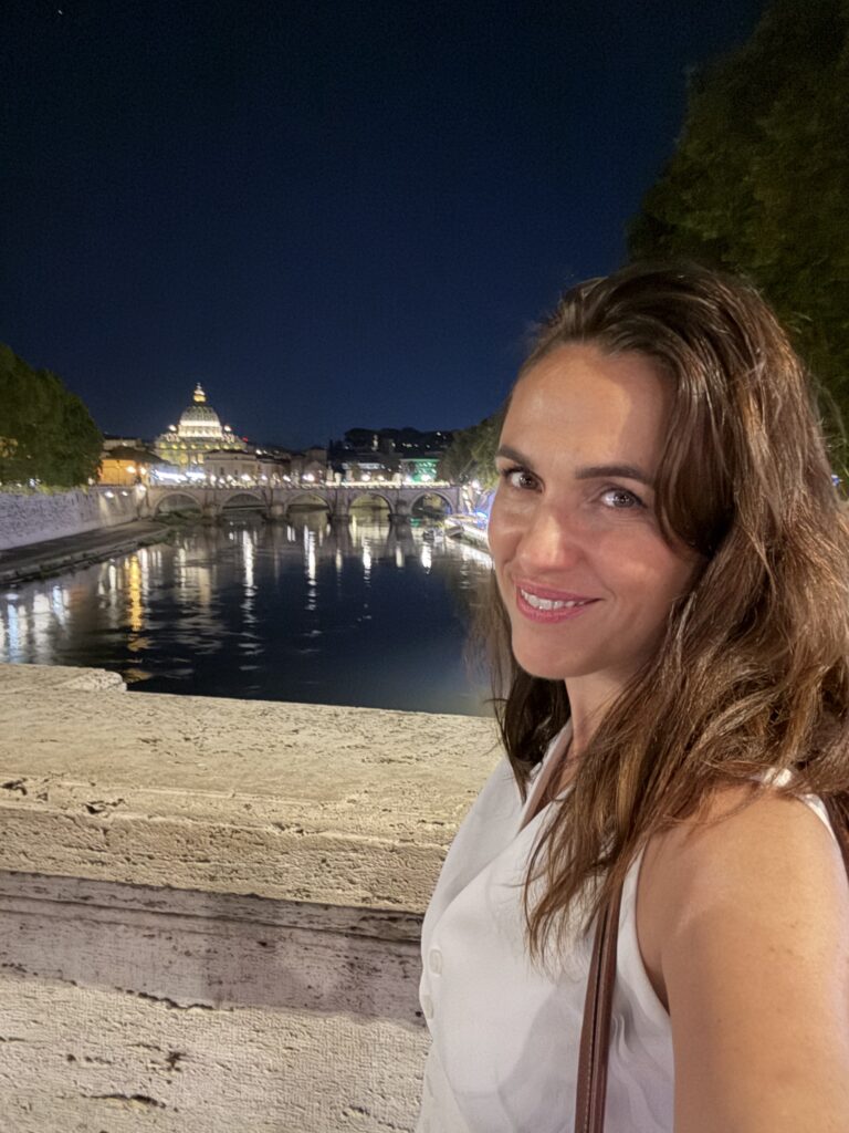 Žena pozira noću uz rijeku, s osvijetljenim gradskim znamenitostima u pozadini.